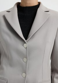 Blazer beige ajusté avec trois boutons métalliques ronds porté sur un pull noir à col rond texturé.