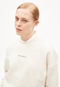 Crème kleurige sweatshirt met hoge halslijn, geborduurd "ARMEDANGELS" logo in beige, zachte textuur, relaxte pasvorm en zichtbare naaddetails.