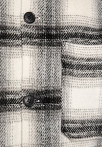 Tissu à carreaux noir et blanc avec une texture douce, présentant un bouton noir et un détail de poche cousue sur un fond clair.