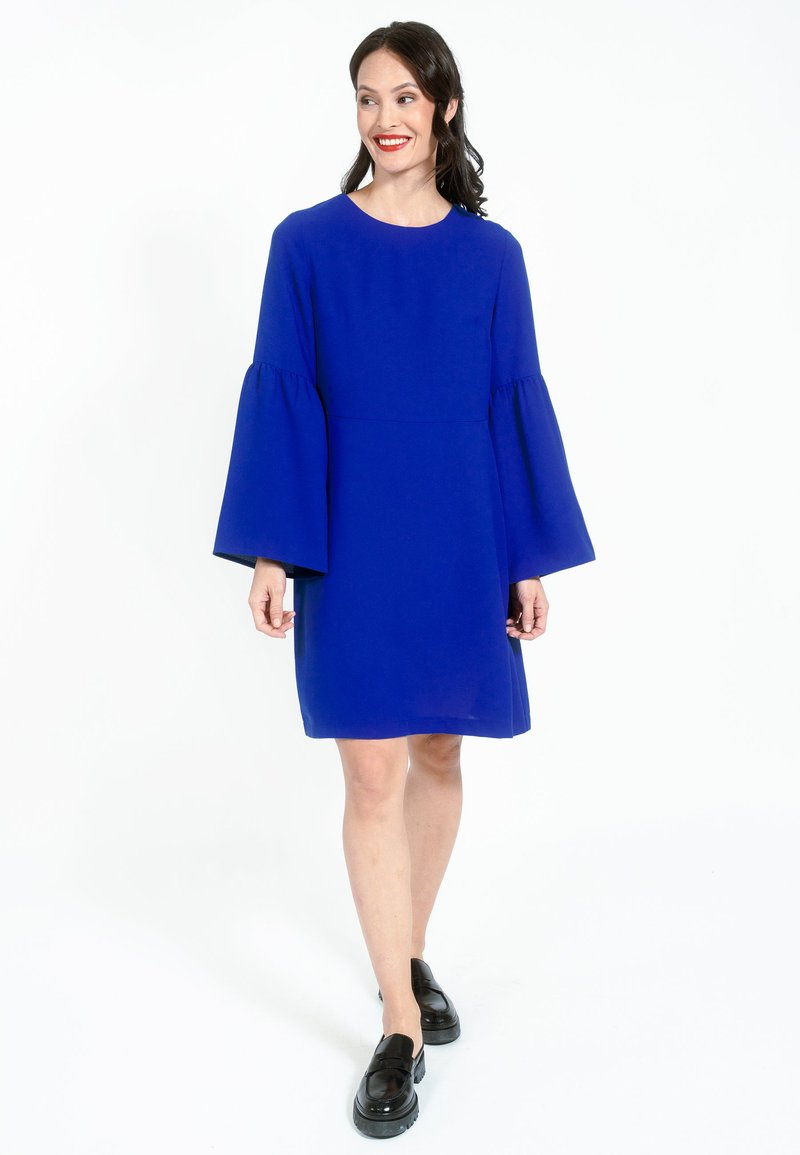Rochie lungă albastră cu mâneci clopot, decolteu rotund și tiv până la genunchi. Asortată cu pantofi cu platformă negri. Textură de țesătură netedă.