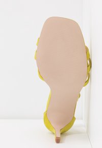 Vue de la semelle d'une sandale jaune à talon haut avec un talon fin et une semelle extérieure lisse beige clair sur fond blanc.