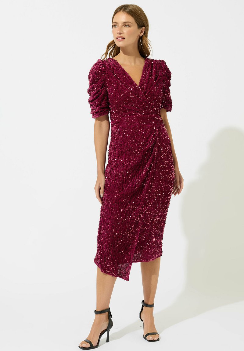 Robe en sequins bordeaux avec manches bouffantes, devant drapé, coupe mi-longue et une subtile brillance. Portée avec des talons noirs à lanières de cheville.