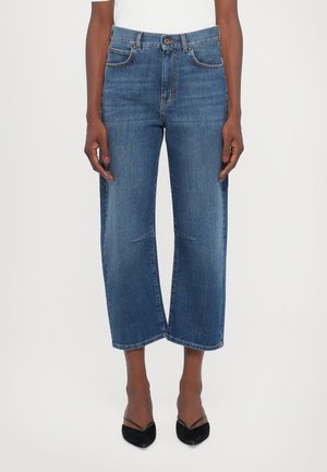 Femme portant un jean large court bleu et des talons noirs pointus, debout, les bras détendus le long du corps, sur fond blanc.