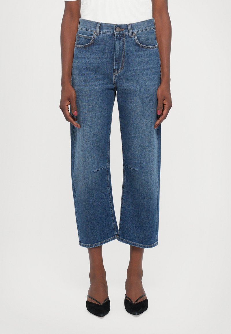 Femme portant un jean large court bleu et des talons noirs pointus, debout, les bras détendus le long du corps, sur fond blanc.