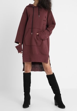 Nainen pukeutuneena viininpunaiseen, ylisuurikokoiseen kulutettuun hupparimekkoon, jossa on etutasku, ja mustiin polven yli ulottuviin korkokenkiin.