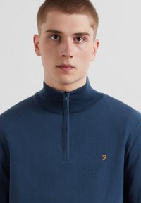 Sweat à fermeture éclair bleu marine avec col montant, poignets côtelés et logo orange brodé sur la poitrine gauche. Texture lisse.