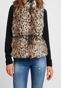 Faux-Fur-Weste mit einem Leopardenmuster in Braun und Creme, ausgestattet mit einem Kragen und einem schwarzen Kordelzuggürtel. Trägt man über einem schwarzen Langarmoberteil.