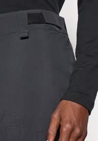 Pilkos, lengvos kelnės turi plokščią juosmenį su Velcro uždarymu ir tiesiu kirpimu; derinamos su ilgomis rankovėmis juoda marškinėliais.