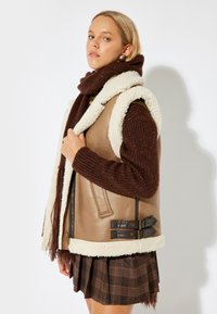 Gilet in pelle color cuoio con imbottitura in shearling bianca, dotato di due fibbie in pelle marrone. Il modello indossa un sciarpa a maglia marrone e una gonna a quadri.