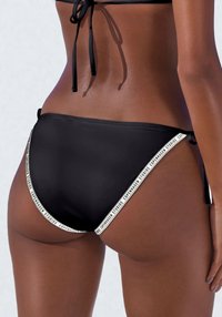 Svart bikinitruse med sidesnøring, med hvit merket kant langs kantene. Glatt tekstur og minimalt dekning design.
