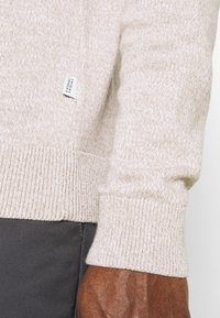 Pull en tricot beige avec poignets et ourlet côtelés, affichant un motif texturé, à côté d'un tissu gris foncé avec des détails de couture visibles.
