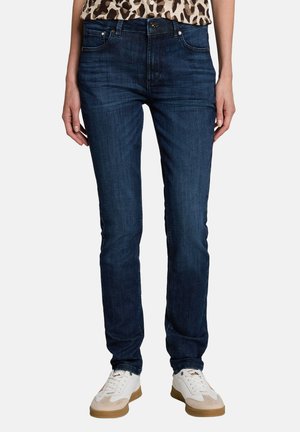 Femme portant un jean bleu foncé, des baskets blanches et un haut à imprimé léopard, debout avec une posture détendue sur un fond uni.