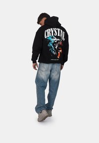 Felpa nera con cappuccio caratterizzata da una grafica colorata di un leone e il testo "CRYSTAL" sul retro; abbinata a jeans baggy azzurri chiari e sneaker grigie.