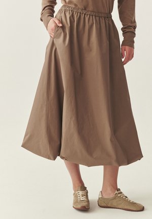 Frau trägt einen braunen Midirock mit elastischem Bund und Taschen, kombiniert mit taupefarbenen Wildledersneakern vor einem schlichten Hintergrund.