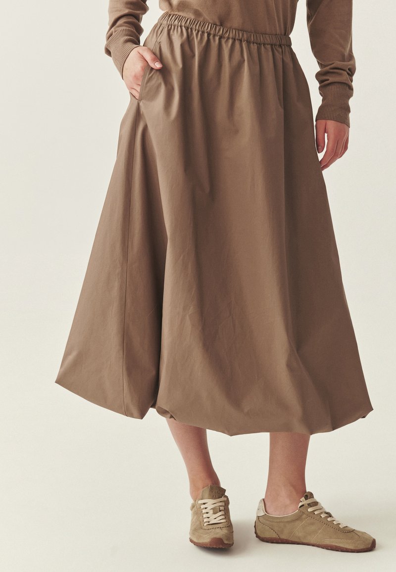 Donna che indossa una gonna midi marrone con vita elastica e tasche, abbinata a sneakers in suede taupe su uno sfondo neutro.