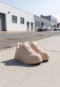 Paire de baskets beiges à plateforme avec des semelles épaisses sur un trottoir urbain ensoleillé à côté d'une rue avec des voitures garées et des bâtiments industriels.