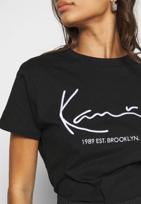 Černé bavlněné tričko s bílým vyšitým textem "KAM" a "1989 EST. BROOKLYN." Kulatý výstřih a krátké rukávy.