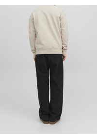 Sudadera beige de manga larga, cuello redondo y dobladillo acanalado, combinada con vaqueros negros de corte holgado y zapatos marrones. Colores sólidos, detalles mínimos.