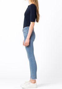 Kvinna som bär en mörkblå topp, ljusblå skinny jeans med trasiga fållar och vita sneakers, stående sidledes på vit bakgrund.