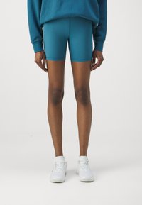 Osoba w dopasowanych turkusowych szortach, turkusowej bluzce z długim rękawem i białych sneakersach stoi na tle czystej białej ściany.