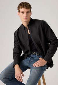 Μαύρο πουκάμισο με κουμπιά από βαμβάκι, φορεμένο πάνω από μια μαύρη φανέλα. Μπλε τζιν παντελόνι με υφασμένη ζώνη. Το μοντέλο κάθεται σε ξύλινο σκαμπό.