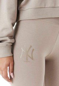 Leggings de sport beiges avec une texture lisse, ornés d'un logo NY doré sur la cuisse et d'un détail de poignet côtelé sur le sweat assorti.