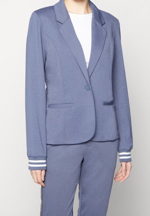 Kobieta ubrana w niebieską dopasowaną marynarkę z jednym guzikiem i spodnie do kompletu, do tego biała koszula pod spodem.