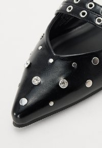 Juodas dirbtinės odos batelis su smailiu varpu, papuoštas sidabriniais spygliais ir blizgučiais, turintis apvalų išpjovą ir plokščią pado.