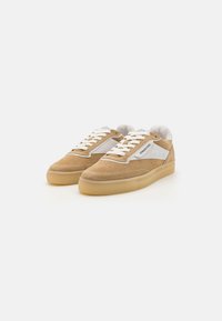 Barna velúr felsőrészű sneaker, fehér bőr díszítésekkel és perforált részletekkel. Kerek orr és átlátszó gumitalp.