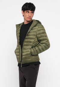 Jacket puffer verde măsliniu cu glugă, închidere cu fermoar și buzunare laterale. Textura ripstop și designul matlasat îmbunătățesc căldura și stilul.