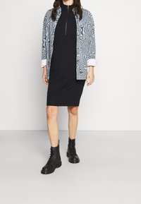 Robe noire avec un col montant sous une chemise grise à motifs, associée à des bottines noires à lacets. Tissu lisse et silhouette ajustée.