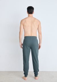 Dunkelgrüne Jogginghosen aus weichem Material mit schmal zulaufendem Bein, elastischem Bündchen und gerippten Abschlüssen, von hinten vor einem hellen Hintergrund präsentiert.