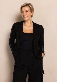 Schwarzer Blazer mit gestreiften Details an den Ärmeln, kombiniert mit einem schwarzen Tanktop und einer Hose. Glatter Stoff, maßgeschneiderte Passform und strukturiertes Design.