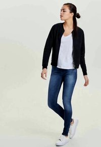 Giubbotto bomber nero sopra una maglietta bianca, abbinato a jeans blu aderenti e sneakers bianche. Outfit casual con design minimalista.