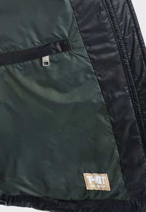 Nahaufnahme eines grünen und schwarzen Jackenstoffs mit einem Reißverschlusstaschen und einem beigen Aufnäher mit der Aufschrift "T-KIT TOUT TEMPS UN".