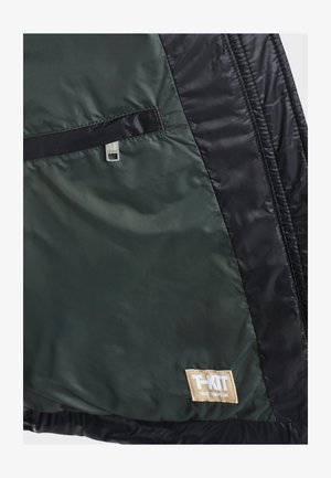 Nahaufnahme eines grünen und schwarzen Jackenstoffs mit einem Reißverschlusstaschen und einem beigen Aufnäher mit der Aufschrift "T-KIT TOUT TEMPS UN".