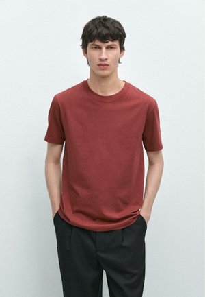 Giovane uomo con capelli corti e scuri, indossa una maglietta a maniche corte rosso mattone semplice e pantaloni neri, in piedi con le mani in tasca.