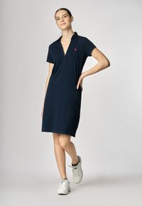 Abito polo blu navy con maniche corte e colletto con zip. Presenta un piccolo logo rosso sul lato sinistro del petto, realizzato in un tessuto morbido e testurizzato.