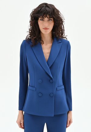 Donna con capelli ricci e scuri che indossa un blazer doppiopetto blu aderente e pantaloni abbinati, in piedi davanti a uno sfondo bianco.
