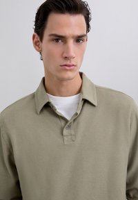 Camisa de polo de manga larga en verde oliva, fabricada en algodón suave, con un cuello, tapeta de dos botones y superpuesta con una camiseta blanca debajo.