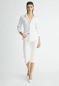 Weißes Hemd mit langen Ärmeln, zwei Brusttaschen; kombiniert mit weißen, cropped Hosen und weißen, spitzen High Heels. Glattes, leichtes Material.
