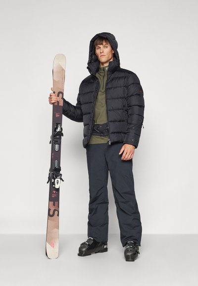 Schwarze Daunenjacke mit Kapuze, olivgrünes Langarmshirt und dunkle Hose. Er hält Skier mit schwarzen Bindungen; Schneestiefel vervollständigen das Outfit.