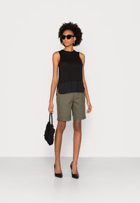 Top preto sem mangas sobre uma saia curta verde-escura, segurando uma bolsa de malha preta. Usa óculos de sol pretos e saltos altos pretos.