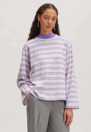 Woman with long curly hair wearing purple-striped long-sleeve shirt, purple turtleneck, gray pants, and dangling earrings, standing against plain background.