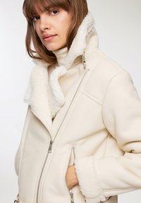 Giacca di pelle color crema con colletto e polsini bianchi sfocati, dotata di zip e tasche laterali in un design strutturato.