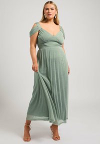 Minttuvihreä iltal dressi, jossa on vartalonmyötäinen yläosa, V-pääntie ja olkaimet ilman olkapäätä. Lattiaan ulottuva, virtaava ja teksturoitu kangas.