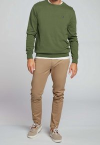 Zelený svetr s kulatým výstřihem vyrobený z pletenin, v kombinaci s hnědými slim-fit kalhotami a světlými teniskami. Jednoduchý design s viditelným švem.