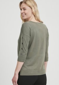 Oluļa zaļš adīts džemperis ar trim ceturtdaļām piedurknēm, ar melnu pogu akcentiem uz piedurknēm un ribota apakšmalu. Gluda tekstūra.