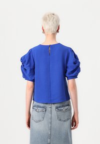 Cobalt blue puff-sleeve top with textured fabric, a round neckline, and a keyhole detail at the back, paired with a denim skirt.