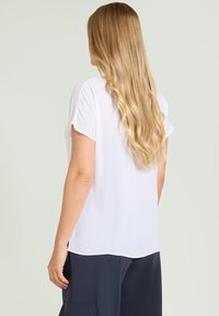 Biała bluzka z krótkimi rękawami, miękki materiał, lekko zaokrąglony dół i rozcięcia po bokach. Model odwrócony plecami, noszący granatowe spodnie.